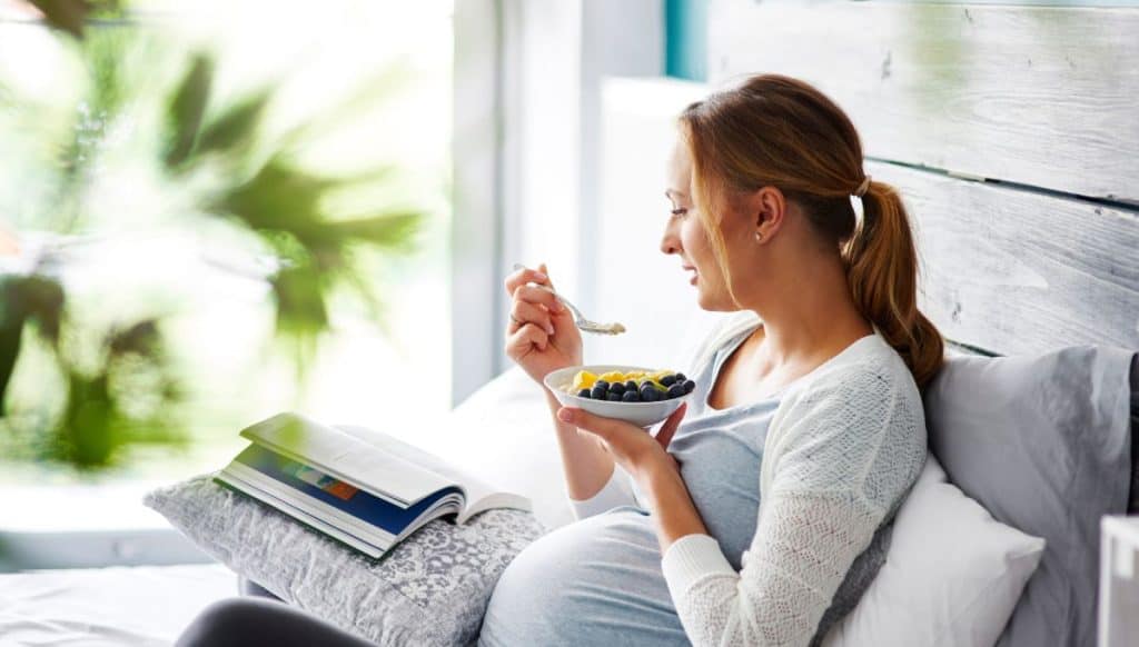 Schwangere sitzt auf der Couch und isst Fruchtmüsli